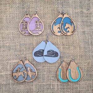 May include: Set of six pairs of wooden teardrop-shaped earrings with various western-themed designs. The designs include cowboy boots, horseshoes, horses, and cowboy hats. The earrings are painted in shades of blue, teal, purple, and brown.