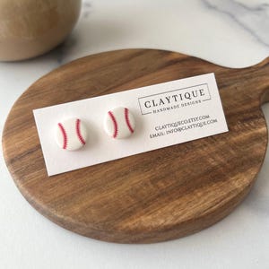 May include: White baseball stud earrings with red stitching, displayed on a white card with the "CLAYTIQUE HANDMADE DESIGNS" logo. The card rests on a wooden cutting board. The earrings are a fun accessory for sports fans.