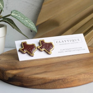 May include: A pair of Texas-shaped stud earrings. The earrings are maroon with a gold-colored outline. They are displayed on a white card with the maker's name and contact information. The earrings are on a wooden cutting board.