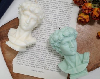 Puede incluir: Dos velas con forma de busto esculpido, una blanca y otra azul claro, sobre una superficie de madera con un libro y flores secas.