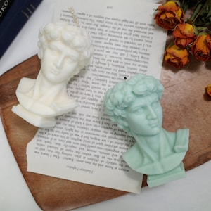 Op de afbeelding: Twee gebeeldhouwde buste kaarsen, een witte en een lichtblauwe, op een houten oppervlak met een boek en gedroogde bloemen.