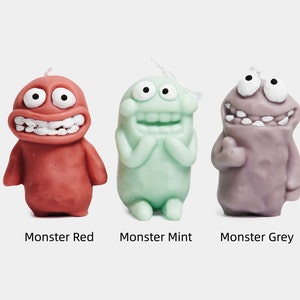 May include: Three monster-shaped candles in red, green, and purple. Each candle has a goofy expression with large eyes and a wide smile. The candles are labeled "Monster Red", "Monster Mint", and "Monster Grey".