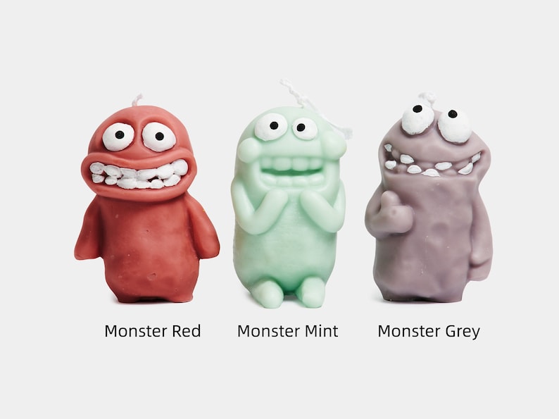 May include: Three monster-shaped candles in red, green, and purple. Each candle has a goofy expression with large eyes and a wide smile. The candles are labeled "Monster Red", "Monster Mint", and "Monster Grey".