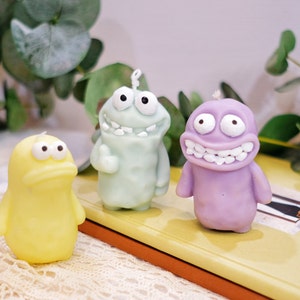May include: Three colorful cartoon character candles. The candles are shaped like green, purple, and yellow creatures with big eyes and toothy grins.