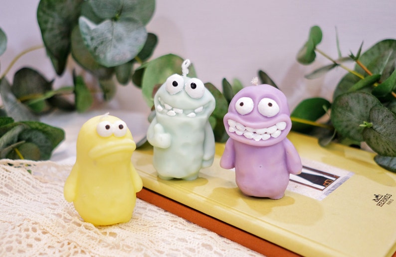 May include: Three colorful cartoon character candles. The candles are shaped like green, purple, and yellow creatures with big eyes and toothy grins.