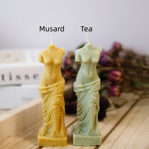 Op de afbeelding: Twee kleine, decoratieve kaarsen in de vorm van het standbeeld van de Venus van Milo. De kaars links is geel en heeft "Venus" op de voet. De kaars rechts is groen en heeft ook "Venus" op de voet. De kaarsen zijn gelabeld met "Musard" en "Tea".