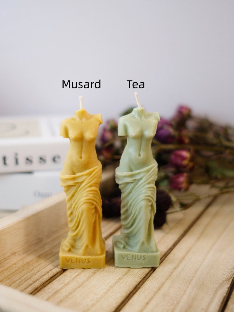 Op de afbeelding: Twee kleine, decoratieve kaarsen in de vorm van het standbeeld van de Venus van Milo. De kaars links is geel en heeft "Venus" op de voet. De kaars rechts is groen en heeft ook "Venus" op de voet. De kaarsen zijn gelabeld met "Musard" en "Tea".