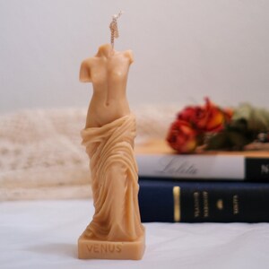 Op de afbeelding: Een beige gebeeldhouwde kaars in de vorm van het standbeeld van de Venus van Milo. De kaars heeft een lont en is op de basis gemarkeerd met "VENUS".