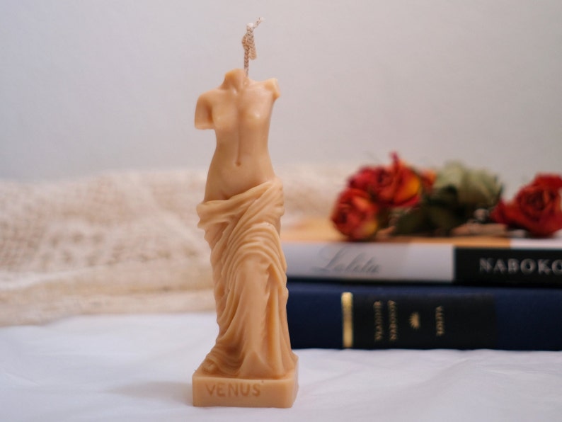 Op de afbeelding: Een beige gebeeldhouwde kaars in de vorm van het standbeeld van de Venus van Milo. De kaars heeft een lont en is op de basis gemarkeerd met "VENUS".