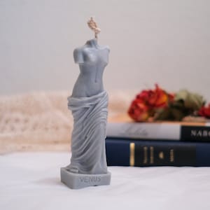 Op de afbeelding: Een grijze gebeeldhouwde kaars in de vorm van het standbeeld van de Venus van Milo. De kaars heeft een lont en staat op een wit oppervlak. De basis van de kaars heeft de inscriptie "VENUS" erop.