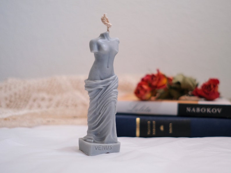 Op de afbeelding: Een grijze gebeeldhouwde kaars in de vorm van het standbeeld van de Venus van Milo. De kaars heeft een lont en staat op een wit oppervlak. De basis van de kaars heeft de inscriptie "VENUS" erop.