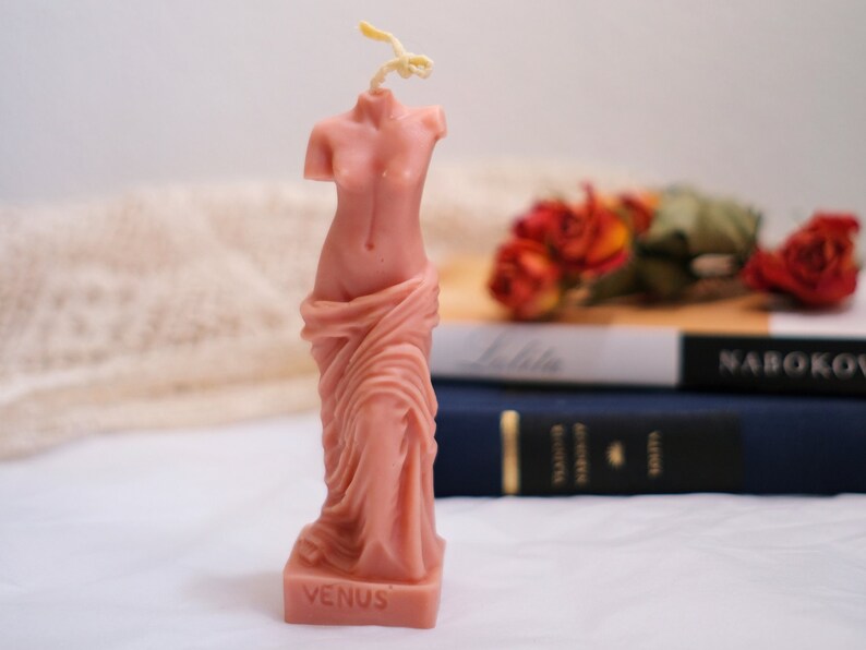 Op de afbeelding: Een roze kaars in de vorm van het standbeeld van de Venus van Milo. De kaars heeft een witte lont en het woord "VENUS" staat op de basis gedrukt.