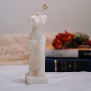 Op de afbeelding: Witte kaars gesculpteerd in de vorm van een gedrapeerd vrouwelijk figuur. De kaars staat op een wit oppervlak met een boek en gedroogde bloemen op de achtergrond.