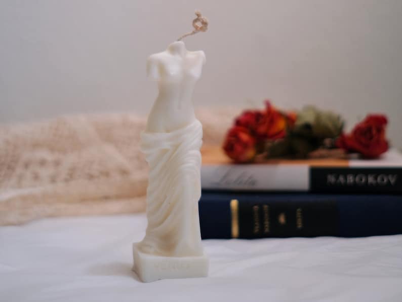 Op de afbeelding: Witte kaars gesculpteerd in de vorm van een gedrapeerd vrouwelijk figuur. De kaars staat op een wit oppervlak met een boek en gedroogde bloemen op de achtergrond.