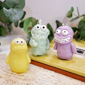 May include: Three colorful cartoon character candles. The candles are green, purple, and yellow. Each candle has a unique facial expression and features large eyes.