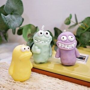 May include: Three colorful cartoon character candles. The candles are shaped like a green, purple, and yellow monster. Each candle has a silly expression on its face.
