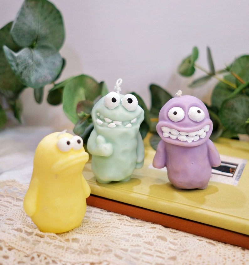 May include: Three colorful cartoon character candles. The candles are shaped like a green, purple, and yellow monster. Each candle has a silly expression on its face.