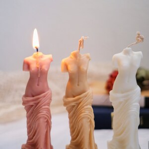 Op de afbeelding: Drie decoratieve kaarsen in de vorm van het standbeeld van de Venus van Milo. De kaarsen zijn roze, beige en wit. De kaarsen hebben het woord "VENUS" op de basis.