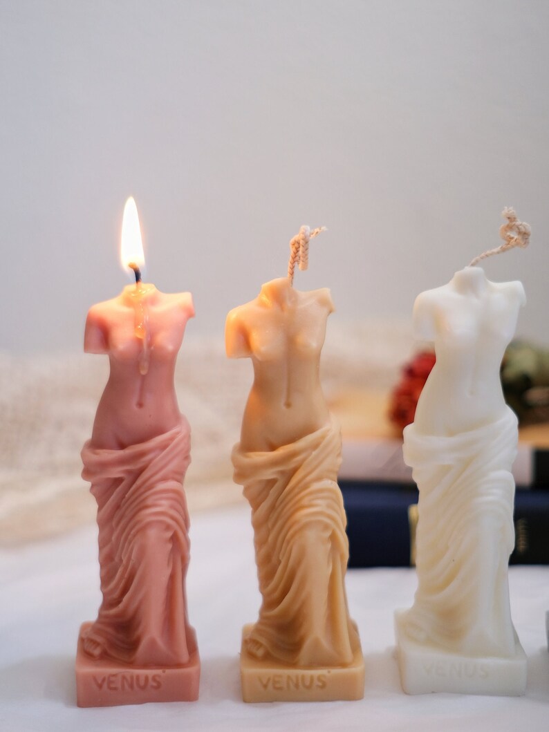Op de afbeelding: Drie decoratieve kaarsen in de vorm van het standbeeld van de Venus van Milo. De kaarsen zijn roze, beige en wit. De kaarsen hebben het woord "VENUS" op de basis.