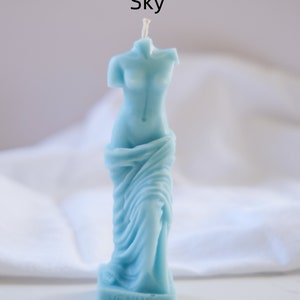 Op de afbeelding: Een lichtblauwe kaars in de vorm van het standbeeld van de Venus van Milo. De kaars heeft een witte lont en de basis van de kaars heeft de tekst "VENUS".