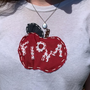 Könnte beinhalten: Weißes T-Shirt mit einem großen, roten Apfel-Applikation. Der Apfel hat den Namen "FIONA" in weißen, genähten Buchstaben. Er hat ein schwarzes Blatt und einen braunen Stiel. Das T-Shirt ist ein lässiges Kleidungsstück für den Alltag.