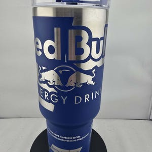 May include: A stainless steel and blue insulated tumbler with the Red Bull logo. The tumbler has a clear lid and is designed to hold 40 fl oz. of liquid. The text "ERGY DRIN" is visible on the tumbler.