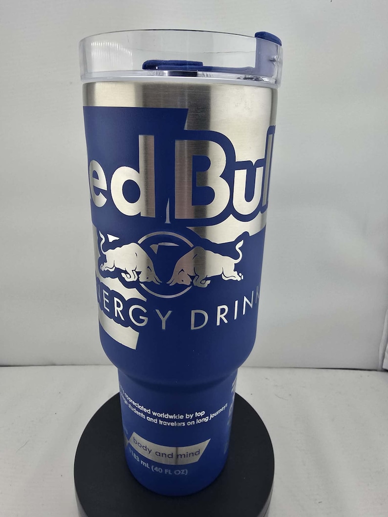 May include: A stainless steel and blue insulated tumbler with the Red Bull logo. The tumbler has a clear lid and is designed to hold 40 fl oz. of liquid. The text "ERGY DRIN" is visible on the tumbler.