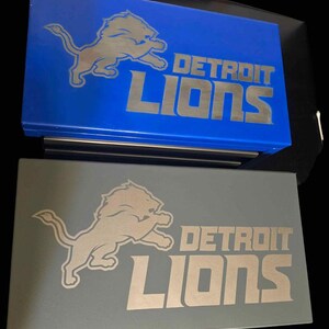 Könnte beinhalten: Zwei Boxen, eine blaue und eine graue, zeigen das Logo und den Text der Detroit Lions in Metallic-Optik. Das Logo ist ein springender Löwe. Der Text lautet "DETROIT LIONS".