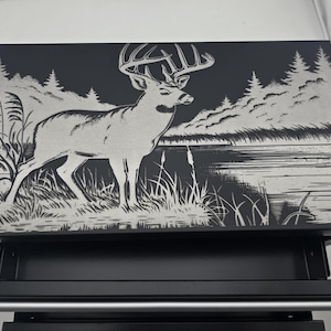 Könnte beinhalten: Schwarze und silberne dekorative Box mit einer detaillierten Illustration eines Hirsches, der in der Nähe eines Sees mit Bäumen im Hintergrund steht. Das Kunstwerk ist auf die Oberseite der Box geätzt und erzeugt einen auffälligen Kontrast.