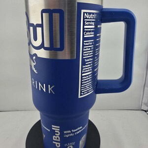 May include: A stainless steel and blue Red Bull drink tumbler with a handle. The tumbler features the Red Bull logo and nutritional information. The tumbler has a clear lid and is designed to keep drinks cold.