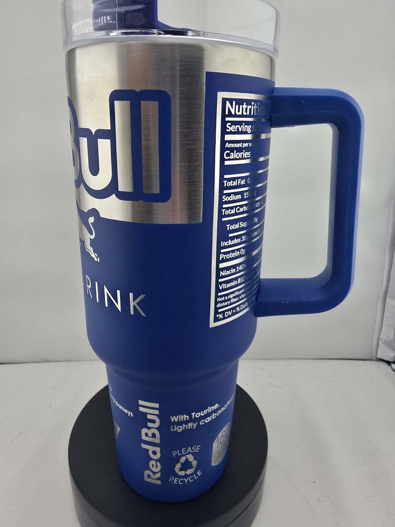 May include: A stainless steel and blue Red Bull drink tumbler with a handle. The tumbler features the Red Bull logo and nutritional information. The tumbler has a clear lid and is designed to keep drinks cold.
