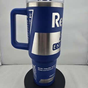May include: A blue insulated tumbler with a handle, featuring the Red Bull logo in white. The tumbler has a clear lid and a silver band. It has a capacity of 1183 ml, and is designed for athletes and busy professionals.