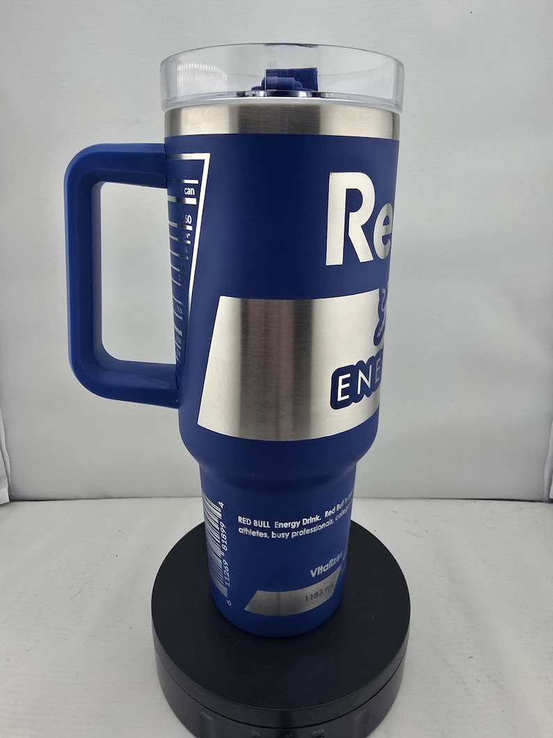 May include: A blue insulated tumbler with a handle, featuring the Red Bull logo in white. The tumbler has a clear lid and a silver band. It has a capacity of 1183 ml, and is designed for athletes and busy professionals.
