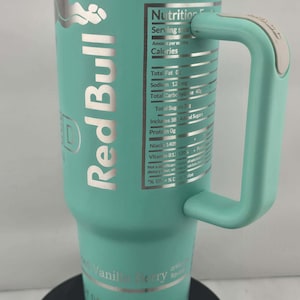 May include: A teal Red Bull travel mug with a handle. The mug features the Red Bull logo and nutrition facts. The bottom of the mug reads "Vanilla Berry" and "160 Calories Per Can".