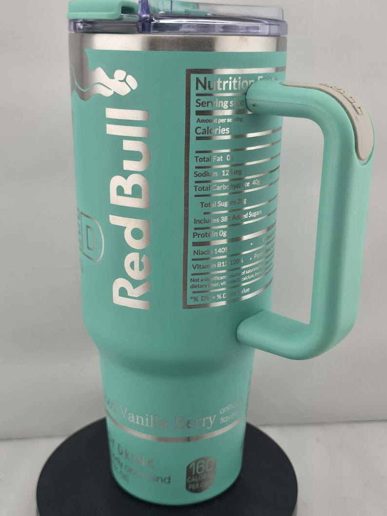 May include: A teal Red Bull travel mug with a handle. The mug features the Red Bull logo and nutrition facts. The bottom of the mug reads "Vanilla Berry" and "160 Calories Per Can".