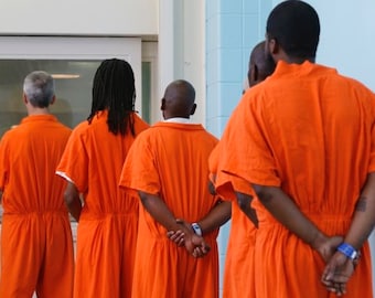 Könnte beinhalten: Fünf Personen in orangefarbenen Overalls stehen in einer Reihe und blicken von der Kamera weg. Sie haben ihre Hände hinter dem Rücken verschränkt.