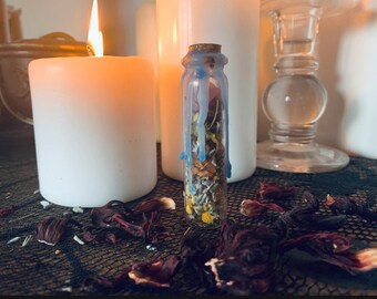 Puede incluir: Un pequeño frasco de vidrio lleno de flores y hierbas secas, sellado con un tapón de corcho. El frasco está parcialmente cubierto de cera azul, creando un efecto de goteo. El frasco está sentado sobre una superficie negra con pétalos de flores secas esparcidos a su alrededor.