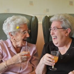 Op de afbeelding: Twee vrouwen zitten op een bank en lachen naar elkaar. Ze houden allebei een glas sinaasappelsap vast. De vrouw links draagt een roze shirt en de vrouw rechts draagt een zwart shirt.