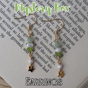 Pode incluir: Brincos pendurados dourados com contas verdes, brancas e pingentes em forma de estrela. Os brincos apresentam uma combinação de contas e pérolas, com um pingente de estrela na parte inferior. A palavra "EARRINGS" está escrita na parte inferior.