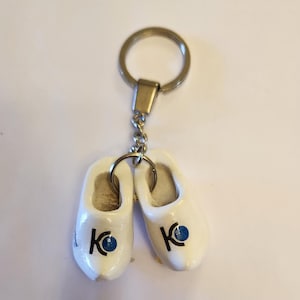 May include: A keychain featuring two miniature white wooden clogs. Each clog has a blue circle with a black "K" on the side. The keychain has a silver-colored metal ring and chain.