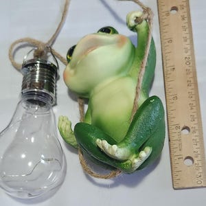 May include: A green frog figurine hanging from a rope, with a clear lightbulb-shaped lamp to the left. The frog has a smiling expression and is holding onto the rope. A ruler is next to the frog. The frog is approximately 4 inches long.