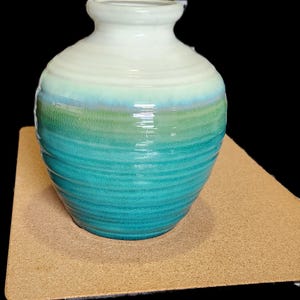 May include: A small, round, ceramic vase with a white top and a teal green bottom. The vase has a textured, ribbed design.