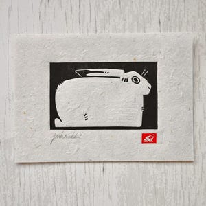 May include: A rectangular piece of textured off-white paper featuring a black and white block print of a rabbit. The print includes the handwritten text jackrabbit in the lower left corner and a small red square stamp with a white icon.