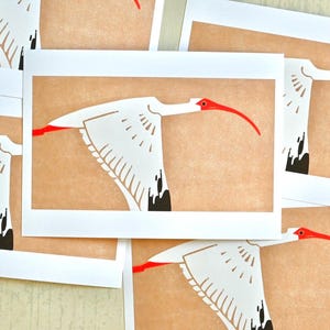 Peut inclure: Plusieurs tirages d'art représentant un ibis blanc en vol. Chaque tirage présente l'oiseau avec un long bec rouge et des pointes d'ailes noires sur un fond beige. Les tirages sont disposés sur une surface de couleur claire.