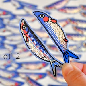 May include: Set of two blue and silver glitter fish stickers. The fish have red and white details on their faces. The stickers are perfect for adding a touch of whimsy to your crafts, journals, or laptops.
