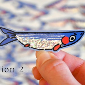 May include: A blue and red fish sticker with glitter on its body. The sticker is on a hand and there are other fish stickers in the background.