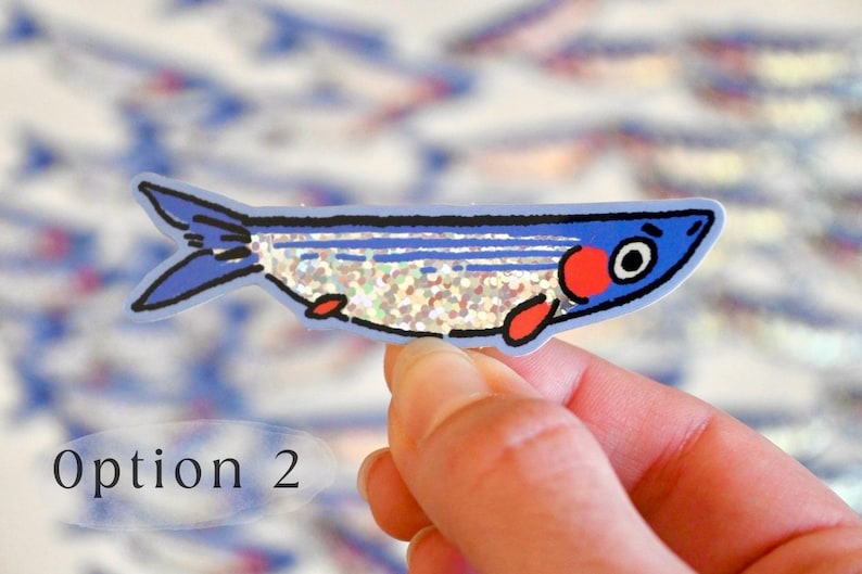 May include: A blue and red fish sticker with glitter on its body. The sticker is on a hand and there are other fish stickers in the background.