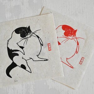 May include: Two black and white prints of a cat on a white background. The cat is depicted in a round, plump shape with its tail curled around its body. The prints are on textured paper and have a red stamp in the bottom right corner.