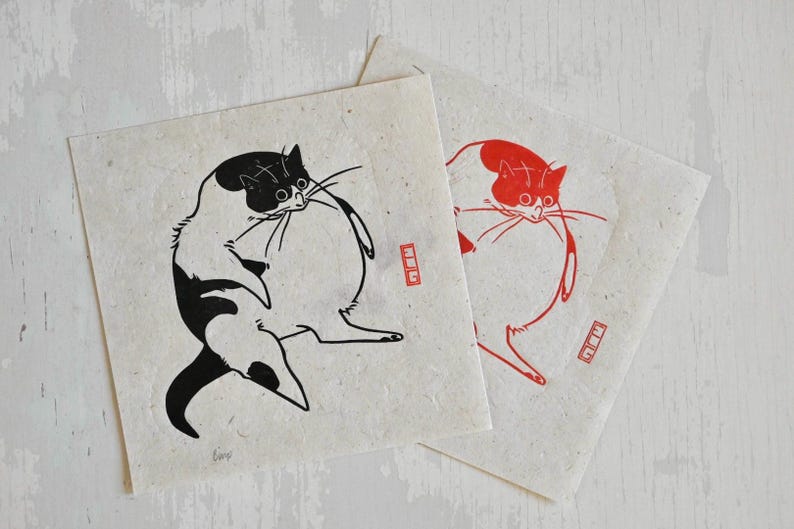 May include: Two black and white prints of a cat on a white background. The cat is depicted in a round, plump shape with its tail curled around its body. The prints are on textured paper and have a red stamp in the bottom right corner.