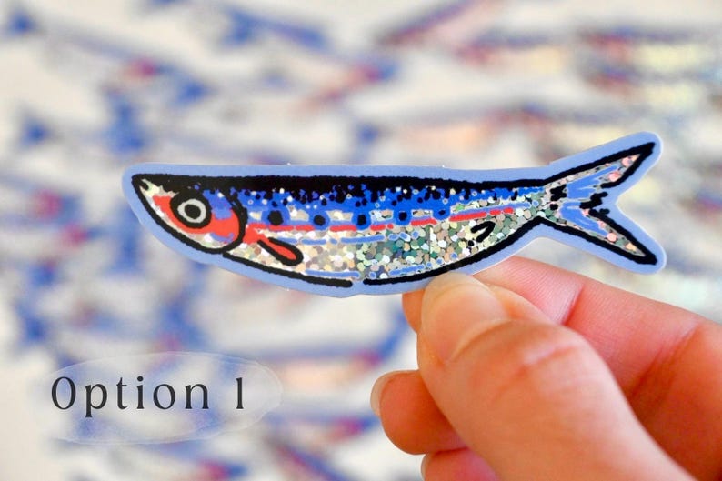 May include: A blue and silver glitter fish sticker with red and white details. The sticker is on a hand.